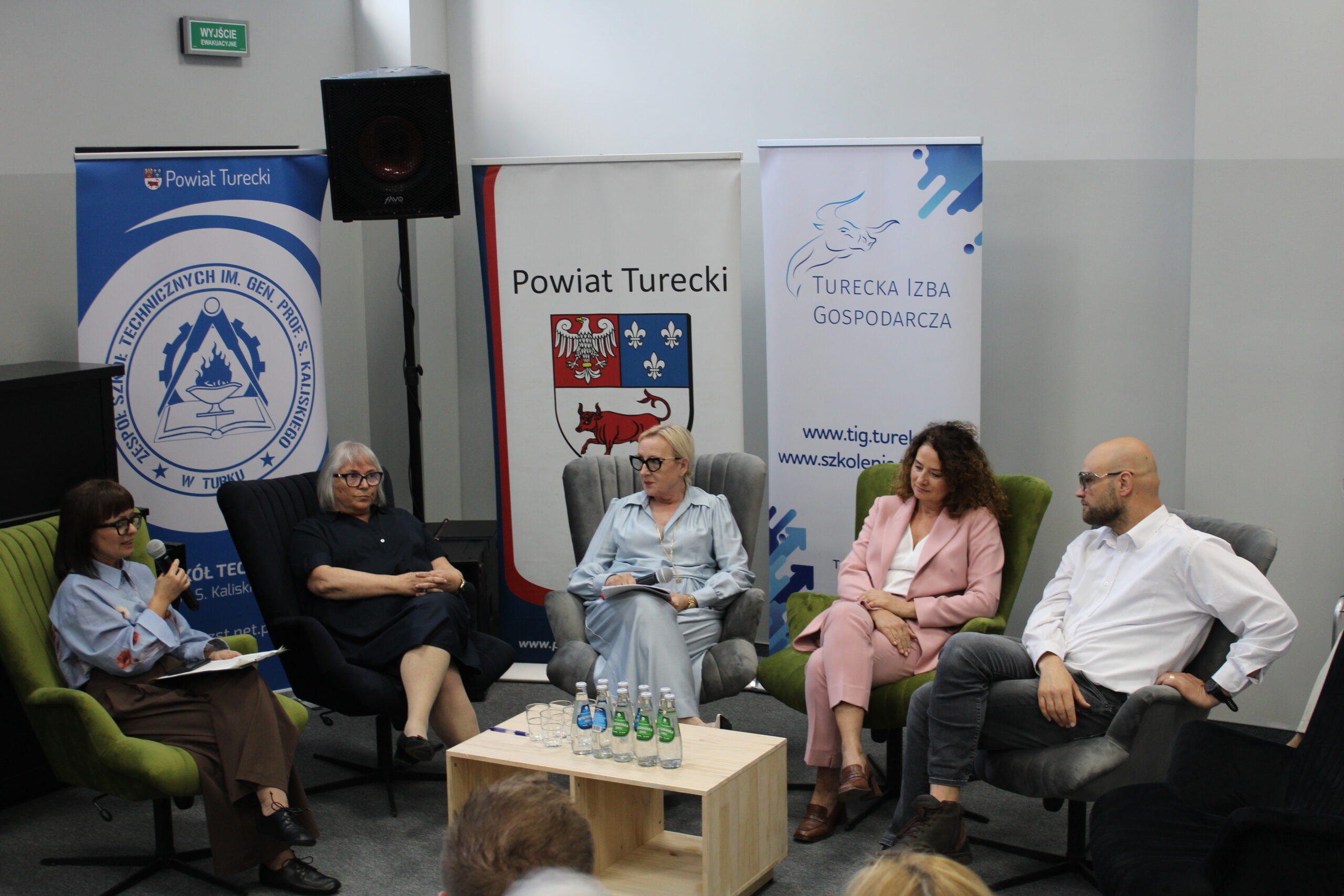 Pięć osób siedzi na krzesłach w panelu dyskusyjnym, obok stoją banery organizacji z powiatu tureckiego.